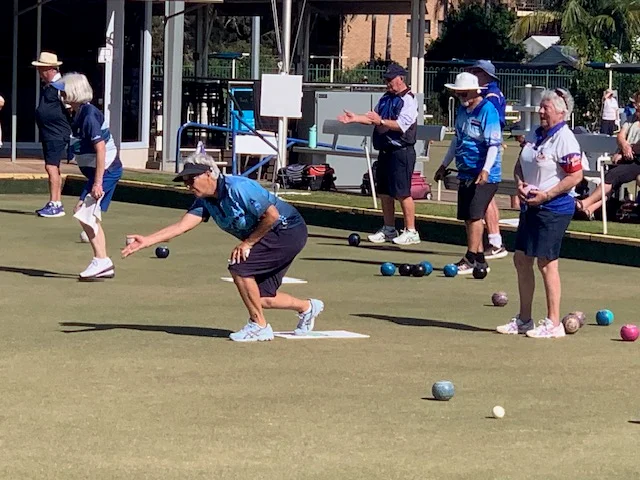 Set on a bowling green, one person almost kneeling, with other bowlers standing behind, and other bowls scattered to the right