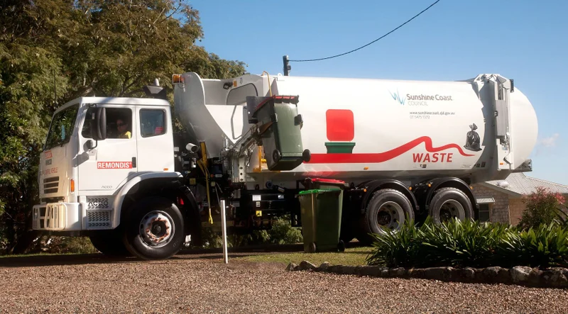 On a residential street, a garbage truck is emptying a red-lidded bin.