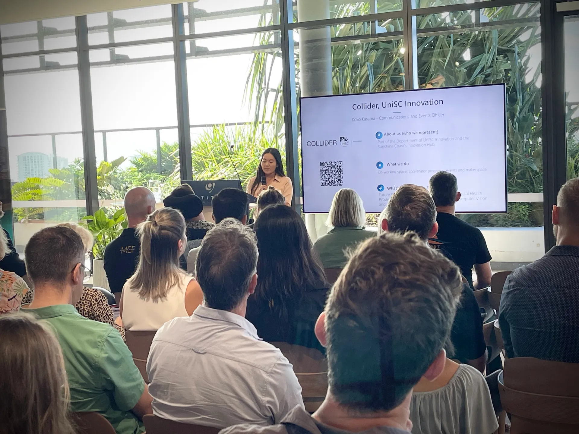 A young woman stands behind a podium talking to a room full of people seated.