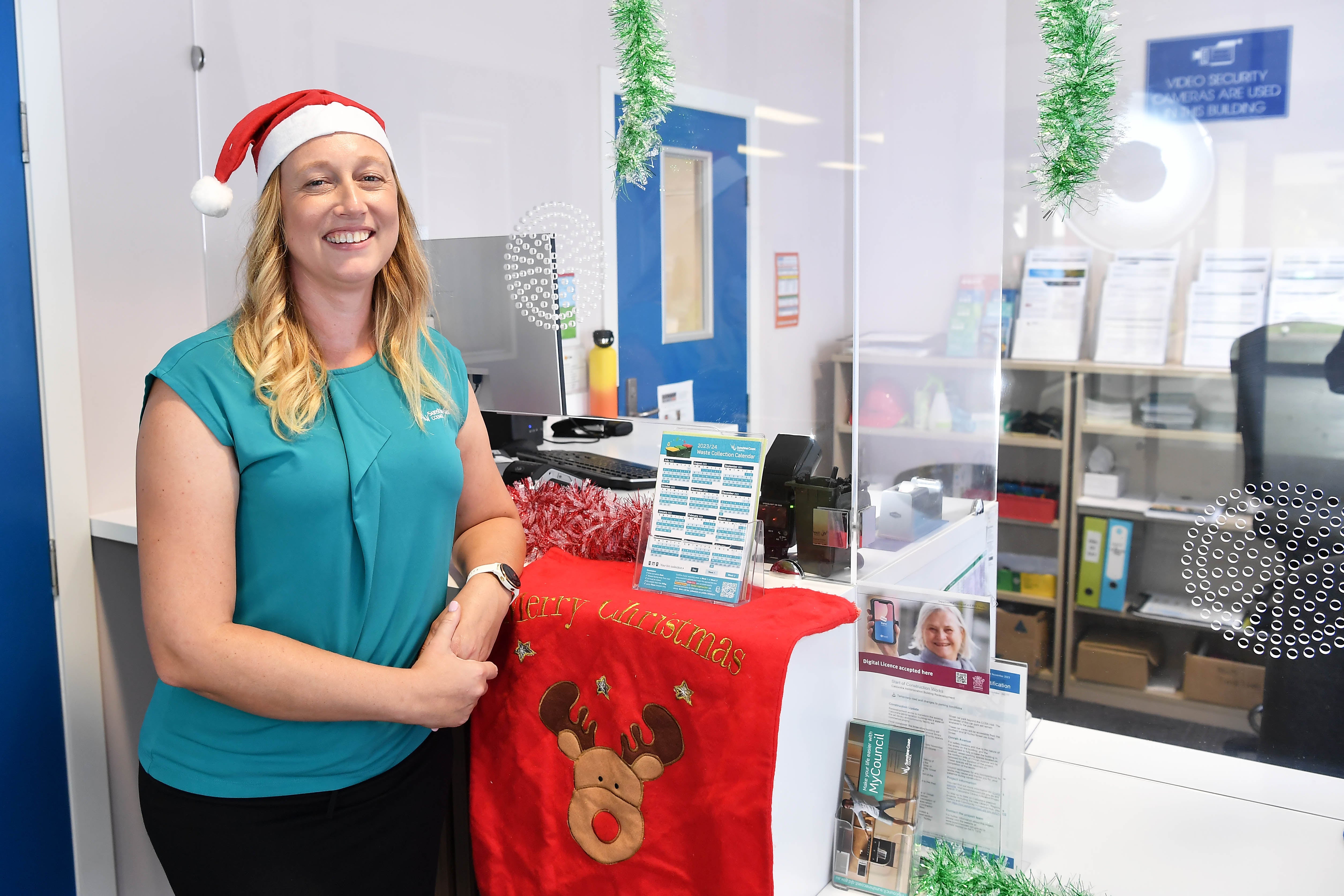 A woman wearing a Santa's hat and a Council uniform leans against a customer service counter that has Christmas decorations on it.