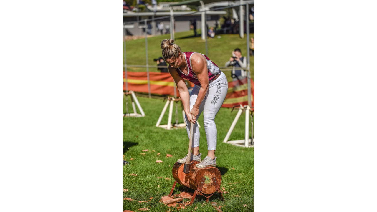 Chopping wood - a lady standing on a wood log bending over it with her axe chopping into the log.