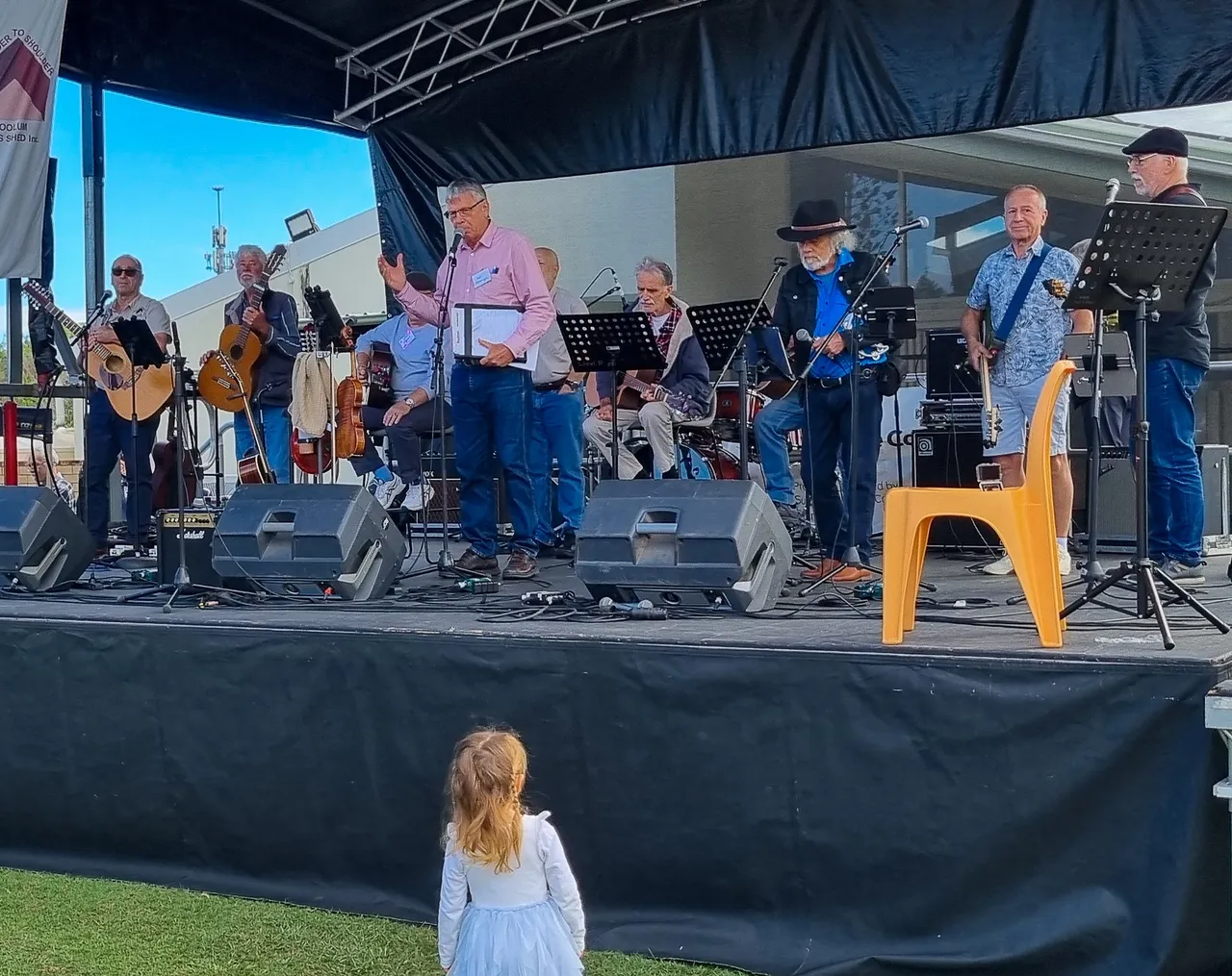 A group of older musicians wait on-stage while a man with a clipboard speaks into a microphone in the stage center. A little girl stands watching at the front.