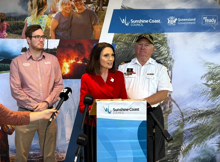 Mayor Rosanna Natoli stands in front of a lectern speaking into four microphones, with two men in uniforms behind her.