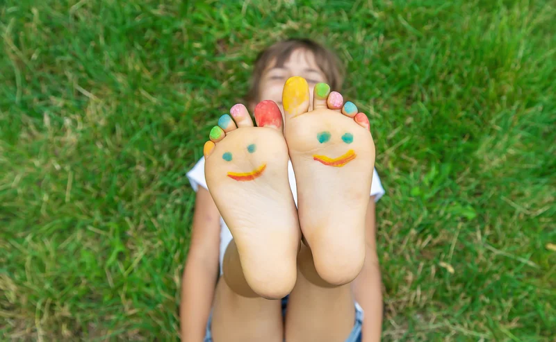 Happy girl lying on green grass with her feet raised up high showing the souls of her feet. Colourful smiley face painted on the souls of her feet.