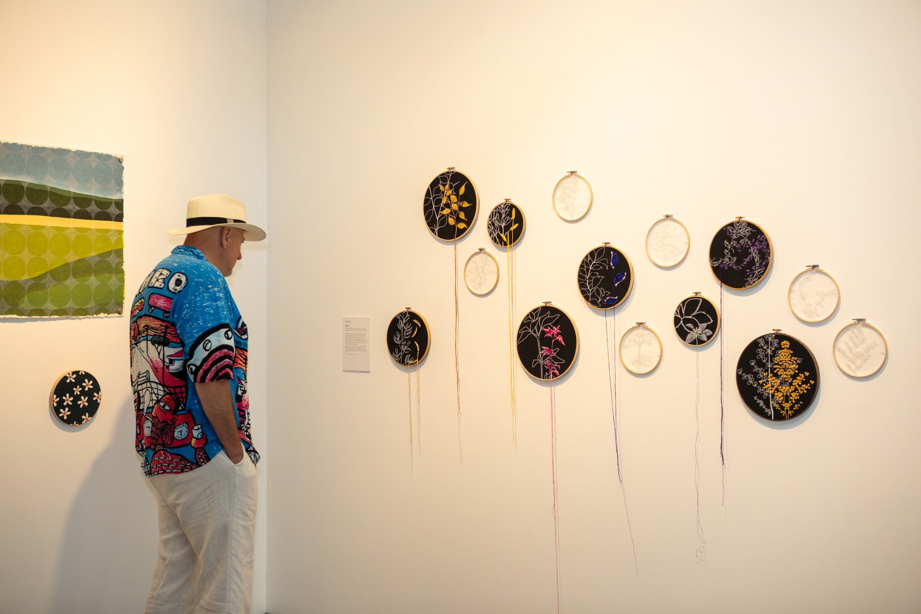 A man in a hat standing in front of a range of artworks at the Wildflowering by Design exhibition launch