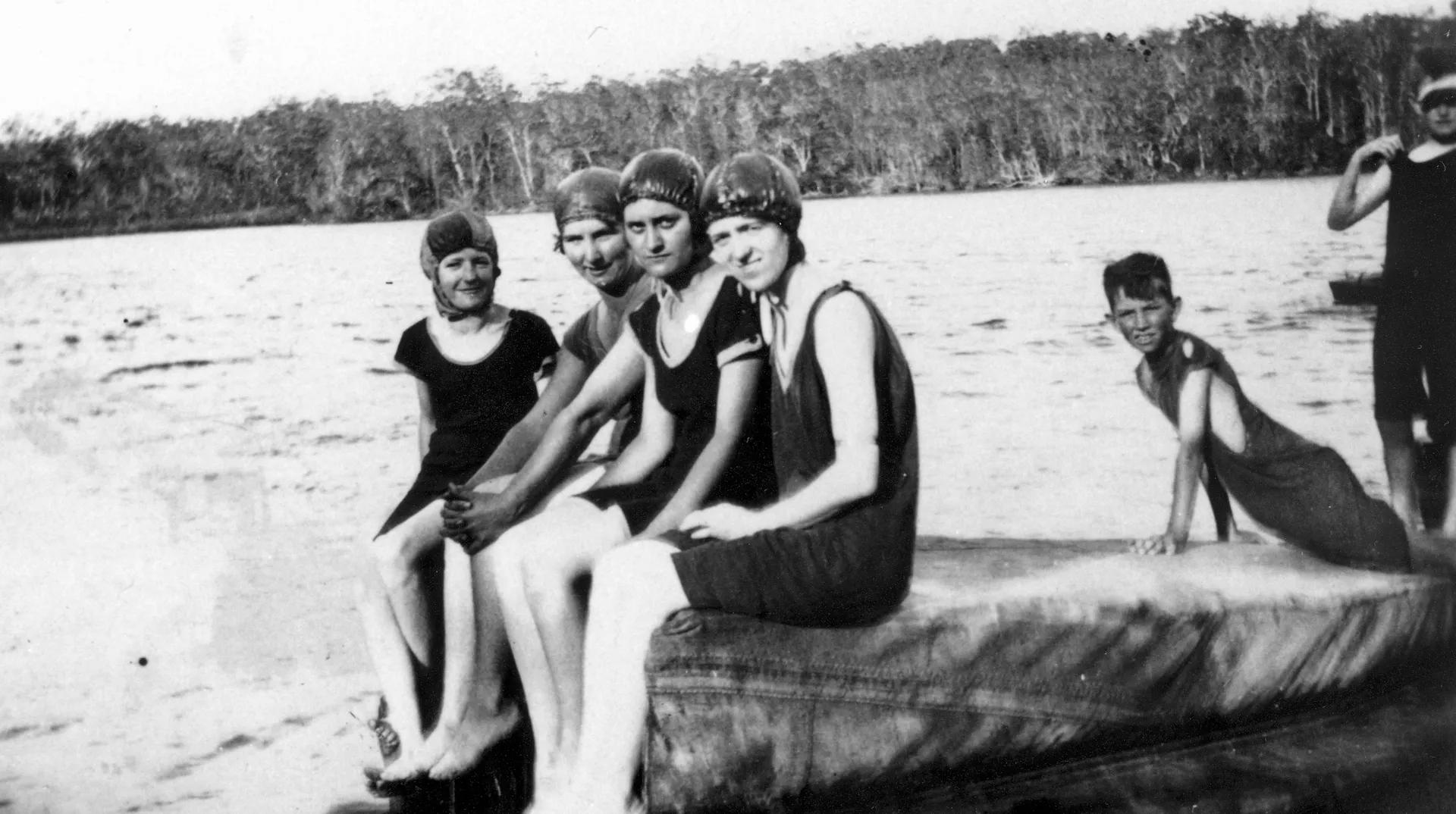 Black and white photo from 1921. Shows four women with swimcaps and dark swim suits sitting on a floating surface on the water. Two young boys lean in for the photo behind them.