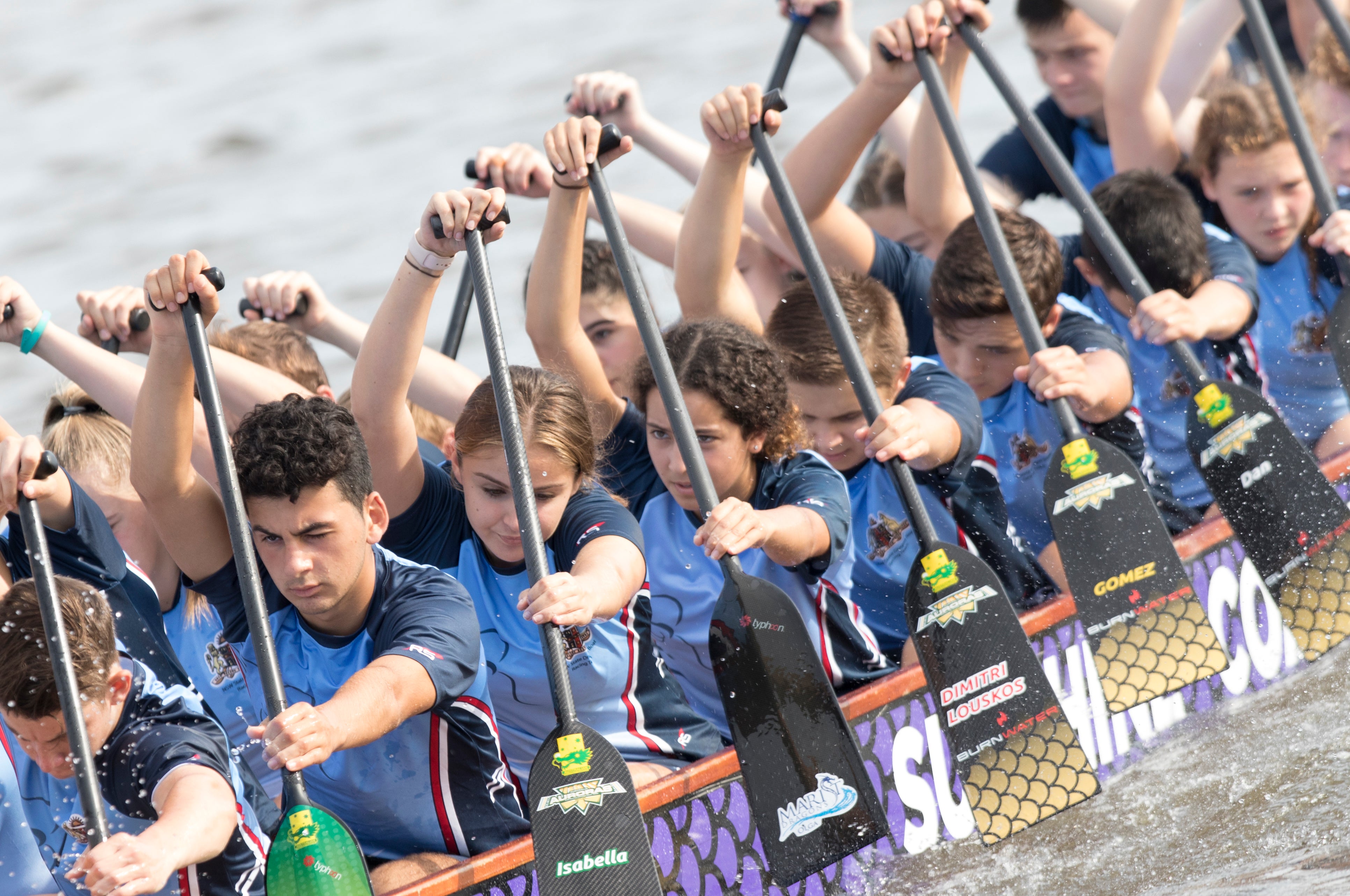 A side photo of a number of Dragon boat teammates with paddles (oars) at the ready to dig into the water.