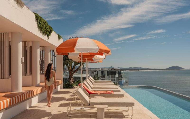 A swimmer walking past orange and seating on the left and sun-lounges with orange and white umbrellas on the deck on her right with the pool in front of those and a sweeping ocean and sky view with Mt Coolum in the background.