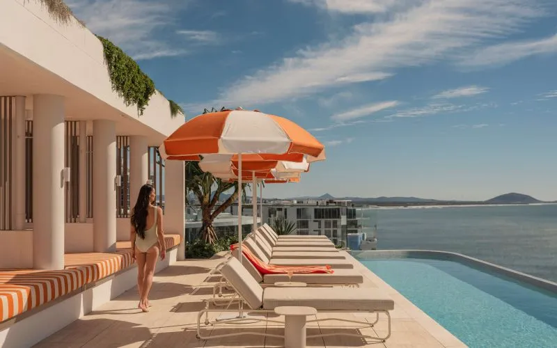 A swimmer walking past orange and seating on the left and sun-lounges with orange and white umbrellas on the deck on her right with the pool in front of those and a sweeping ocean and sky view with Mt Coolum in the background.
