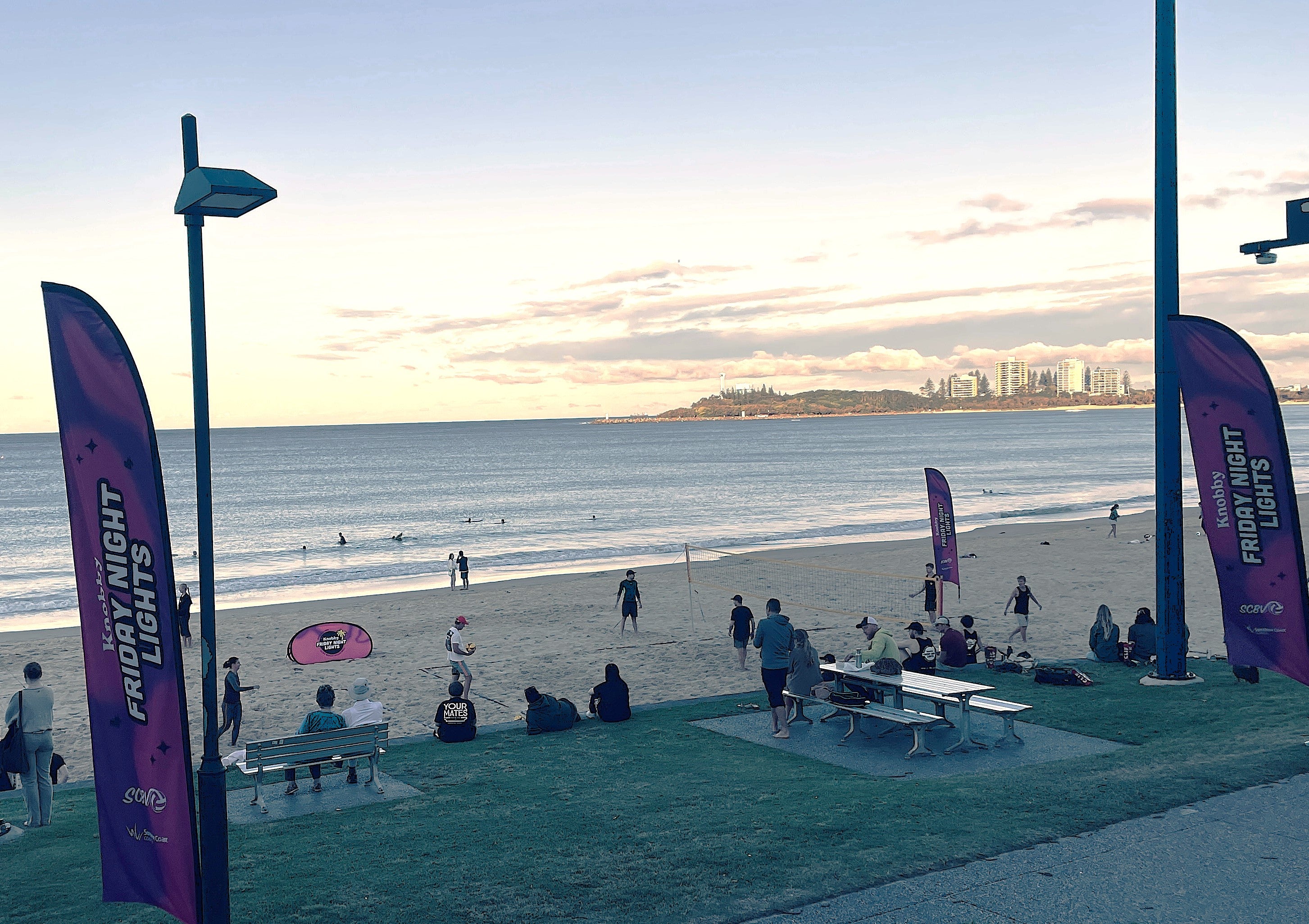 Mooloolaba Beach, volleyball set up on the sand and large purple flags promoting Friday Night Lights set up on the grass overlooking the beach. Crowds of people playing, watching and swimming. 