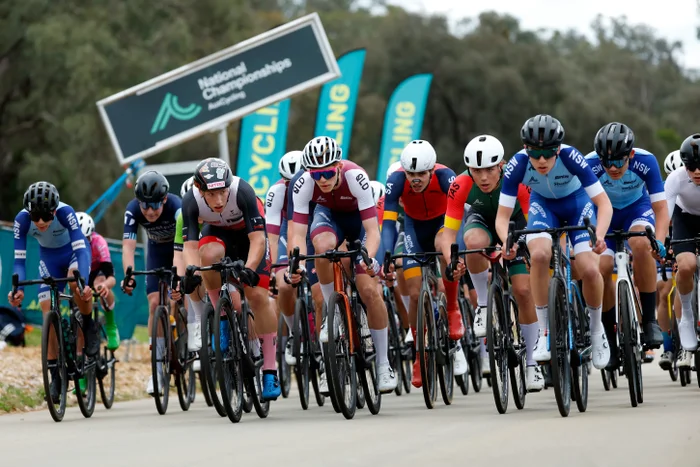 A group of on-road racing cyclists, withe signage in the background that reads National Championships