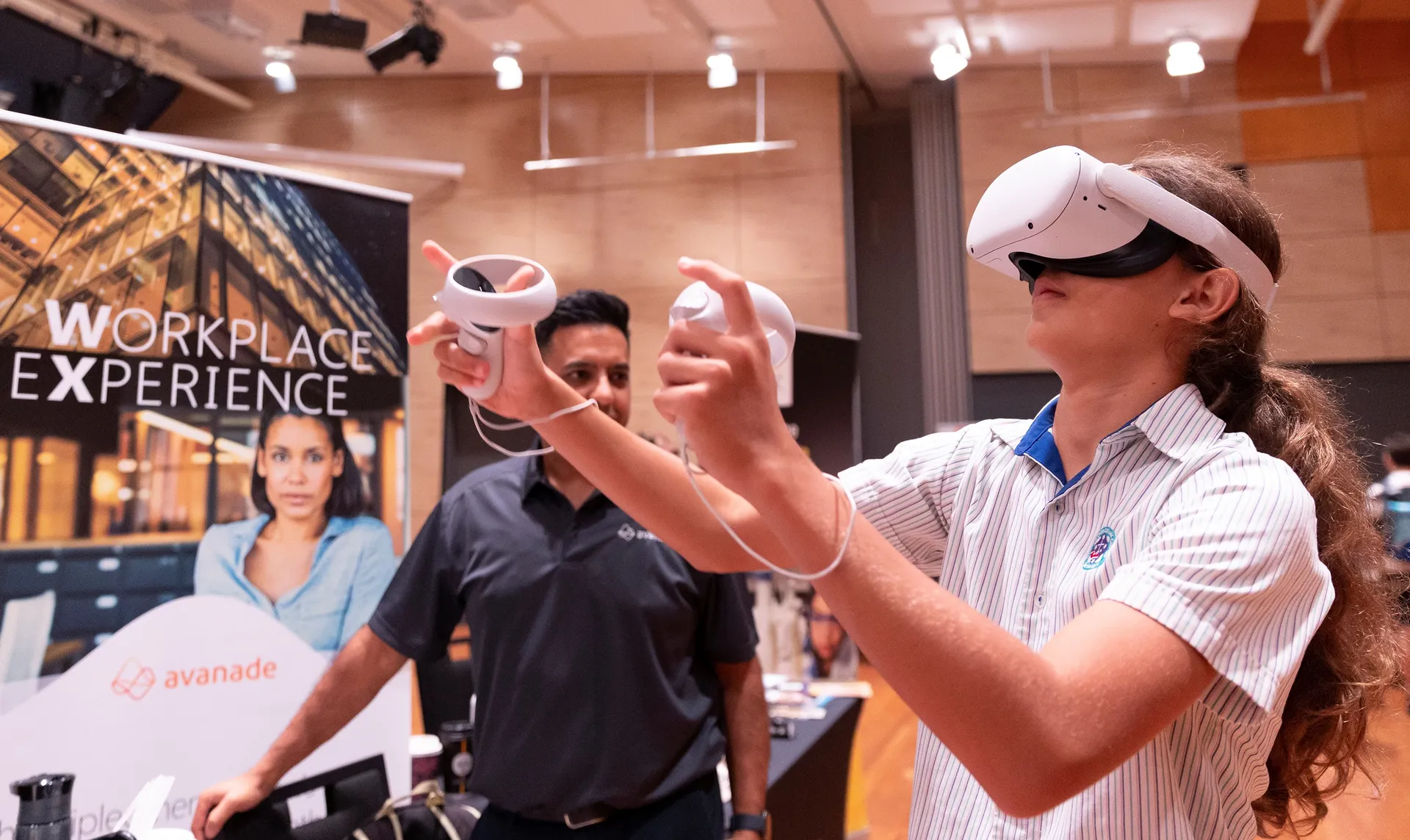 A student trying virtual reality equipment with a supplier next to the student providing guidance. Behind them on the left is a pull up style banner with a person pictured, and the words above their head Workplace Experience.