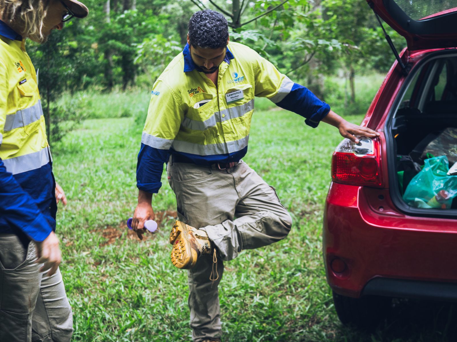A man sprays disinfectant on the soles of his boots, next to a car parked in a forested area.