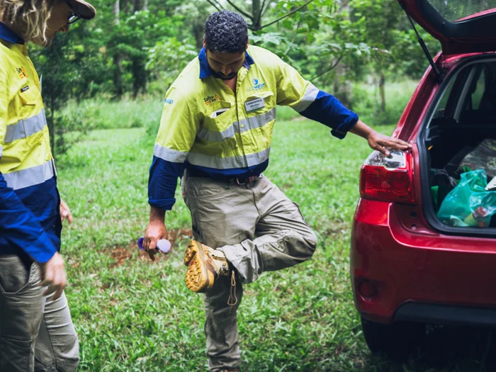 A man sprays disinfectant on the soles of his boots, next to a car parked in a forested area.