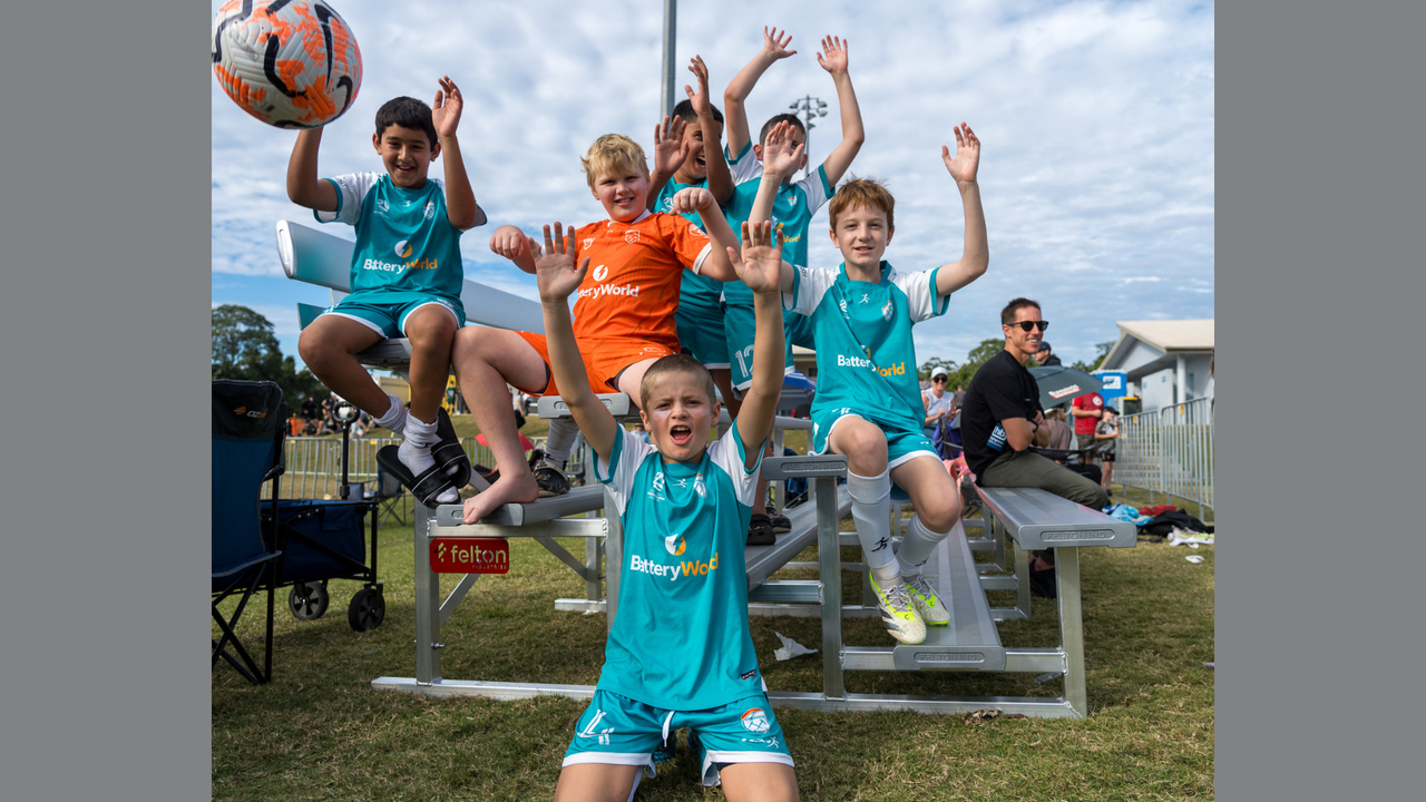 Six young footballers siting on a tiered spectator bench seat with their hands up in the air and one has thrown a soccer ball showing in the top right corner. One player is in a bright orange shirt amongst them, a football referee in the making.