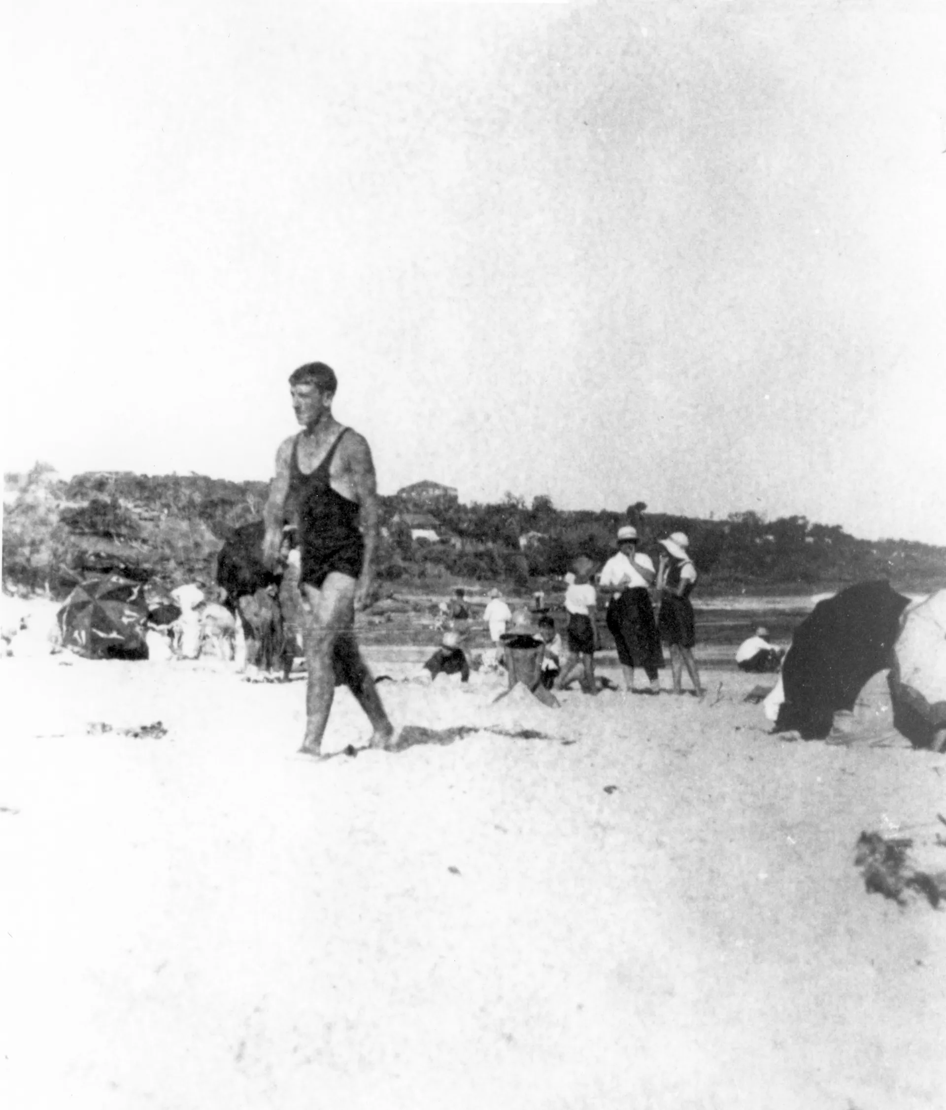 Black and white photo from 1922 of a man in a swimsuit walking on the sand at a busy beach.