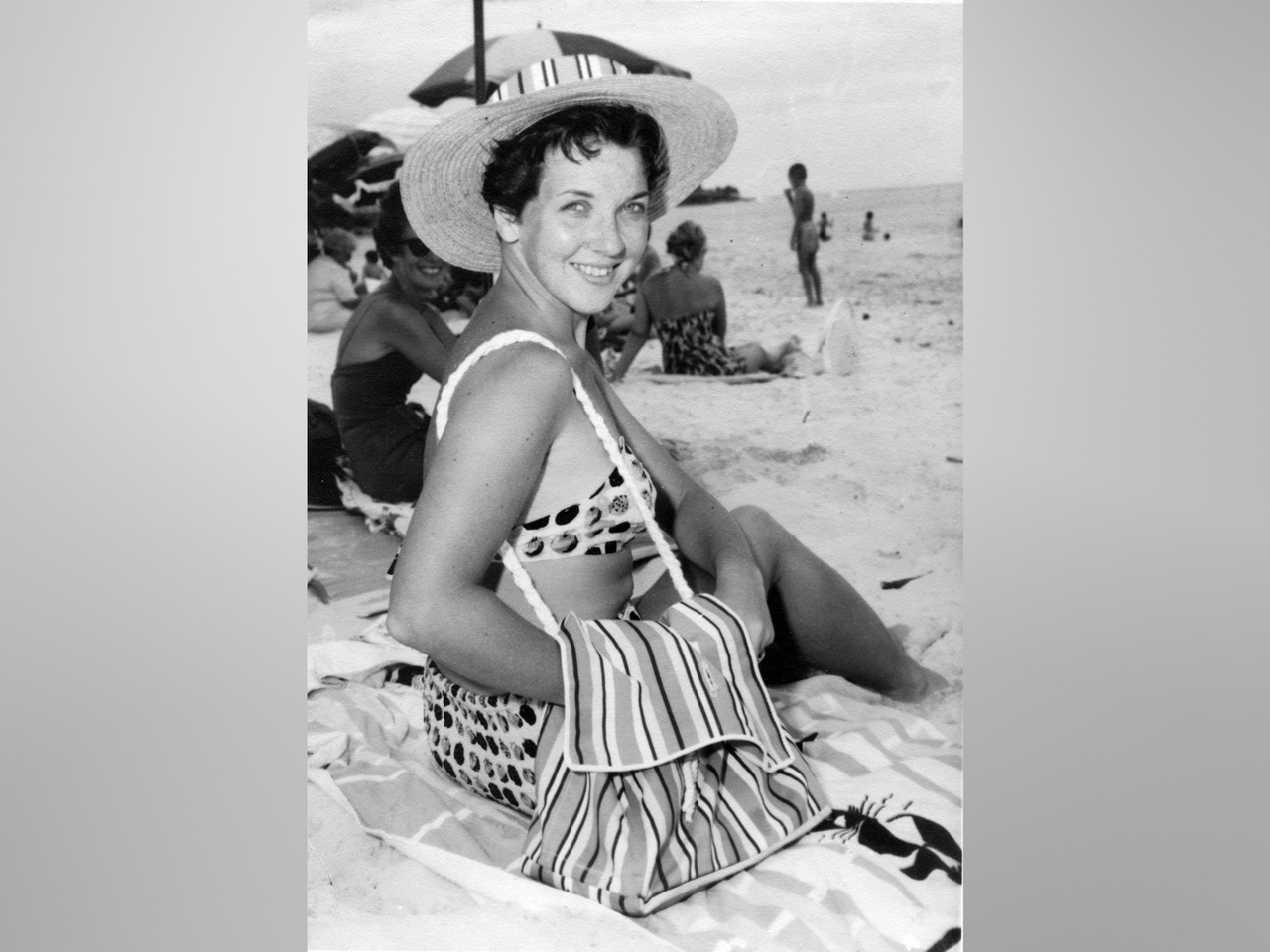 Black and white photo from the 1950s of a smiling woman at the beach with a sun hat and bikini. Bag draped over her.