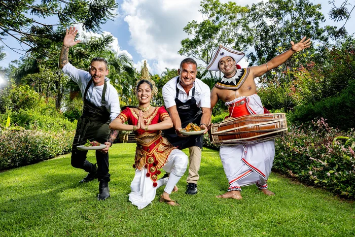 Two chefs each wearing aprons and holding two plates of food; plus two performers - in Sri Lankan garb.