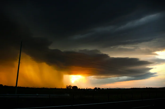 The sun sets behind a dark and menacing clouds as a wall of rain falls on land in silhouette.