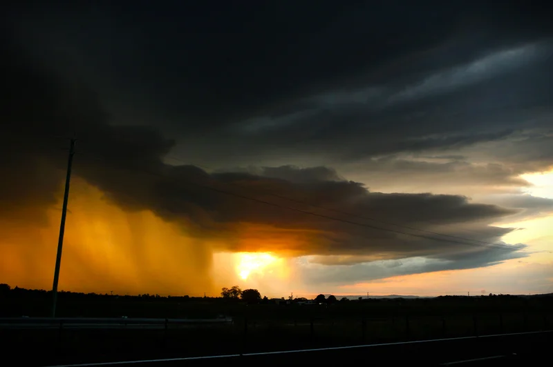 The sun sets behind a dark and menacing clouds as a wall of rain falls on land in silhouette.