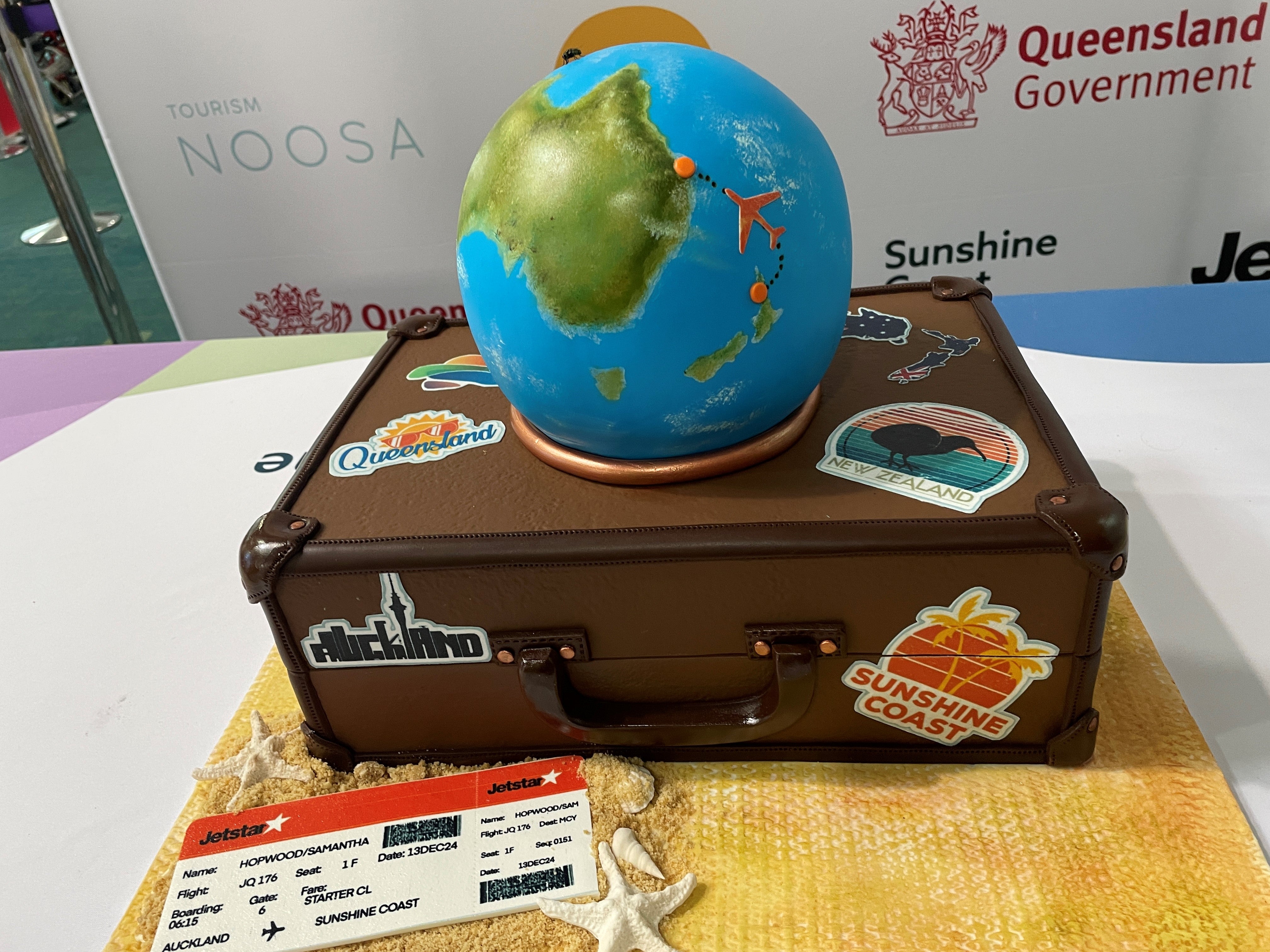 A cake in the shape of an old style brown suitcase with the world globe on top featuring a map of Australia and New Zealand and a Jetstar plane flying between the two countries.