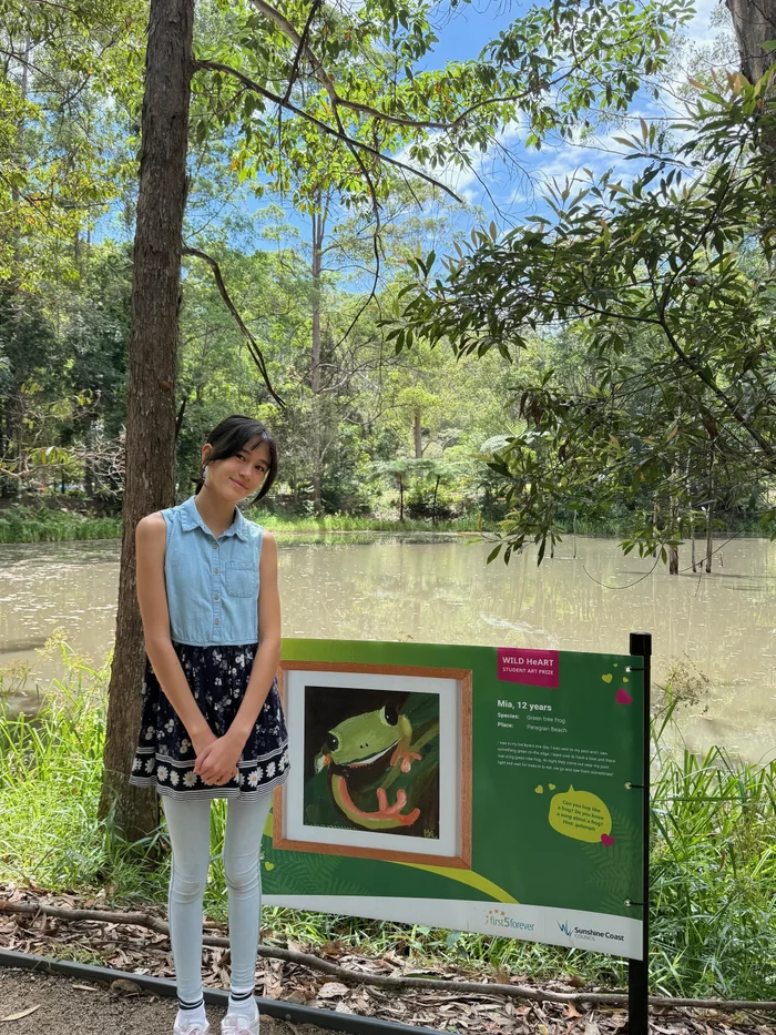 Young girl standing proudly in front of her nature-inspired artwork, displayed beside a tranquil pond at the Maroochy Regional Bushland Botanic Garden during the Wild HeART Student Art Prize exhibition.