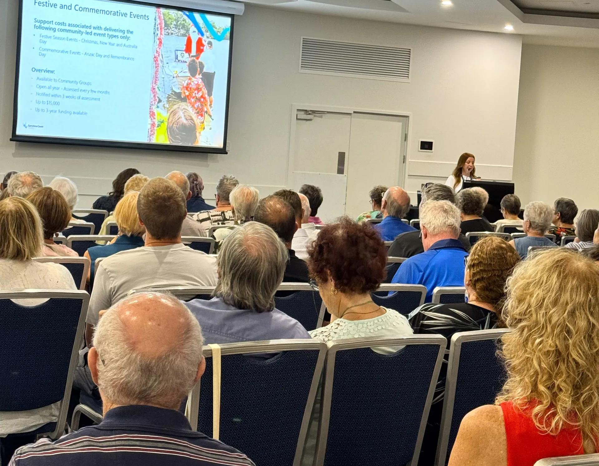 A large group of people are seated in front of a large screen. In the background is a presenter behind a podium.