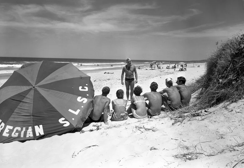 Black and white photo from the 1960s of a man talking to a crowd of beach goers sitting next to a large Peregian S.L.S.C umbrella .
