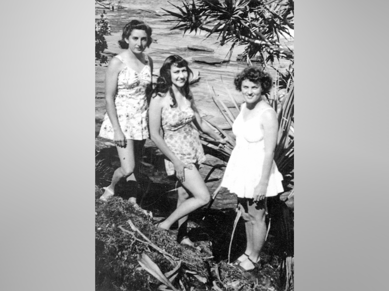 Black and white photo of three women posing in short, strappy dresses at the beach in 1945.