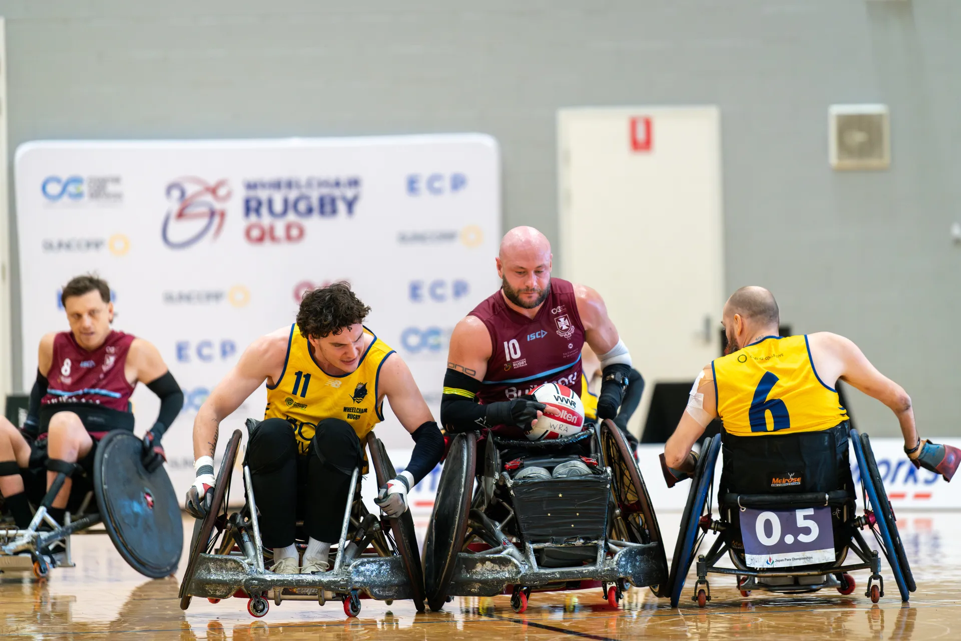 Four parasport athletes - two in yellow bits, two in maroon
2nd on the right is holding the ball and is flanked by the opposing team.