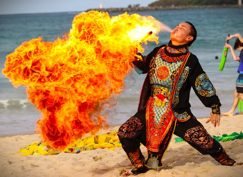 A man on the beach wearing traditional Chinese dress, breathes out a huge ball of fire.