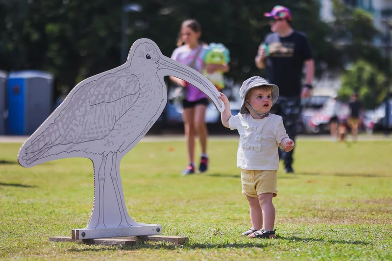 A young child standing with a cut out of a shorebird on green grass in an open park