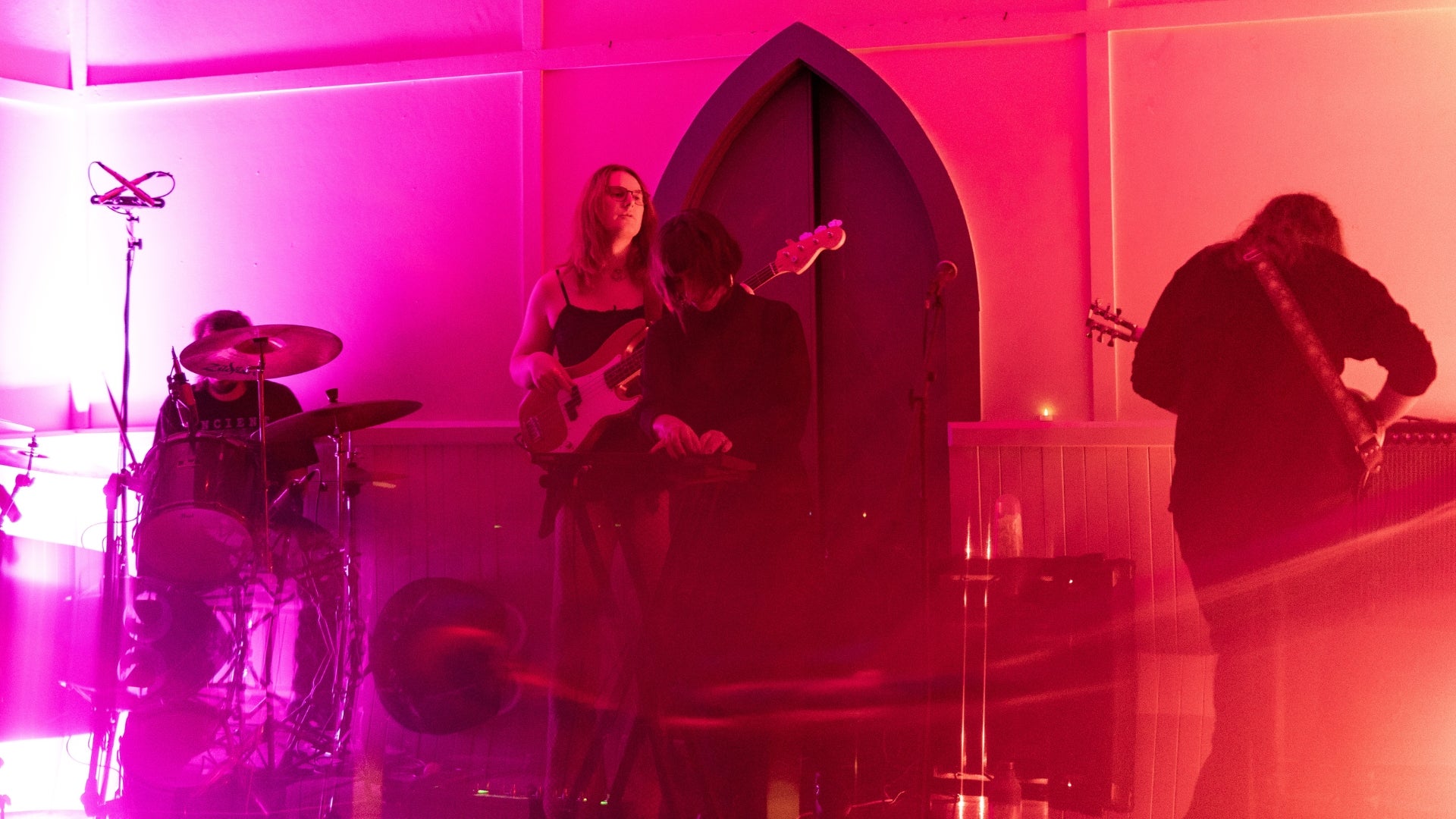 Musicians performing on a hazy, pink and orange lit stage. There is a drummer, two guitar players and someone on keys.