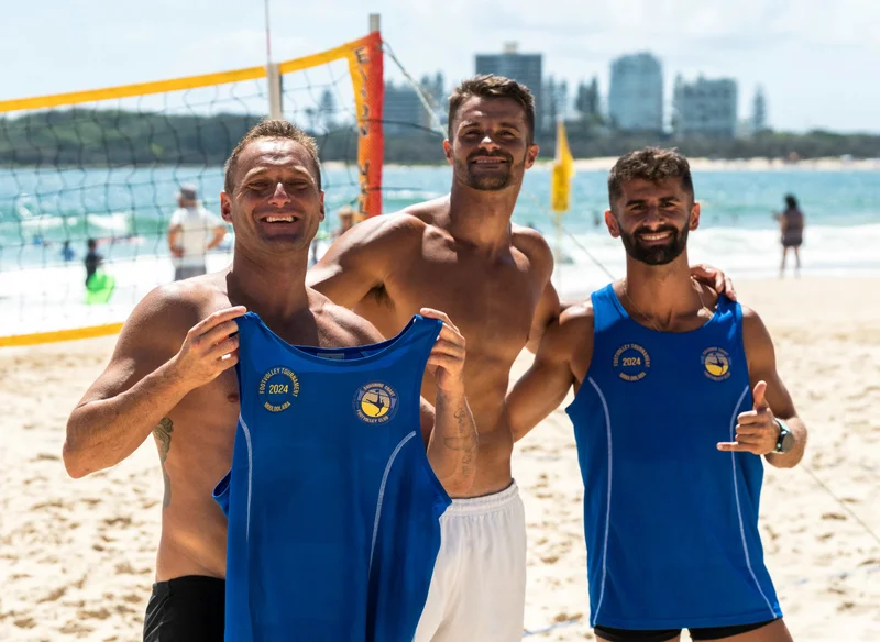 Three smiling men stand in front of a volleyball net on a busy beach in front of a volleyball net, holding up blue team singlets.