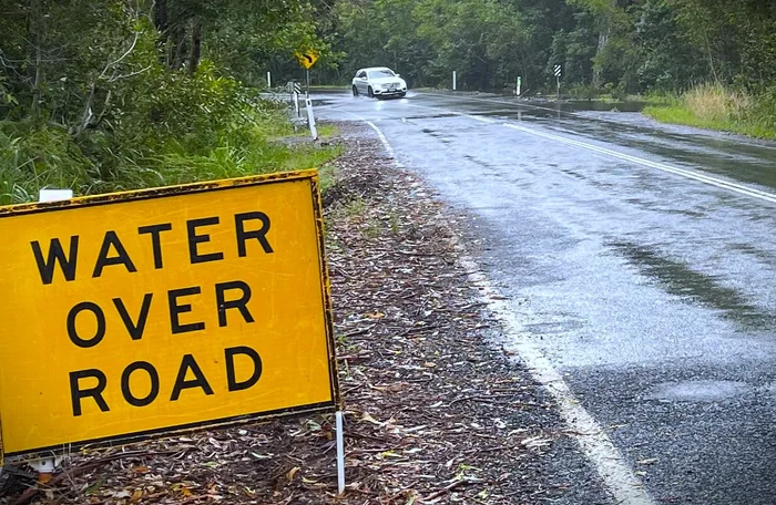 A yellow, water over the road sign sits at the front of the photo on the left. A white car is about to drive into water on the road.