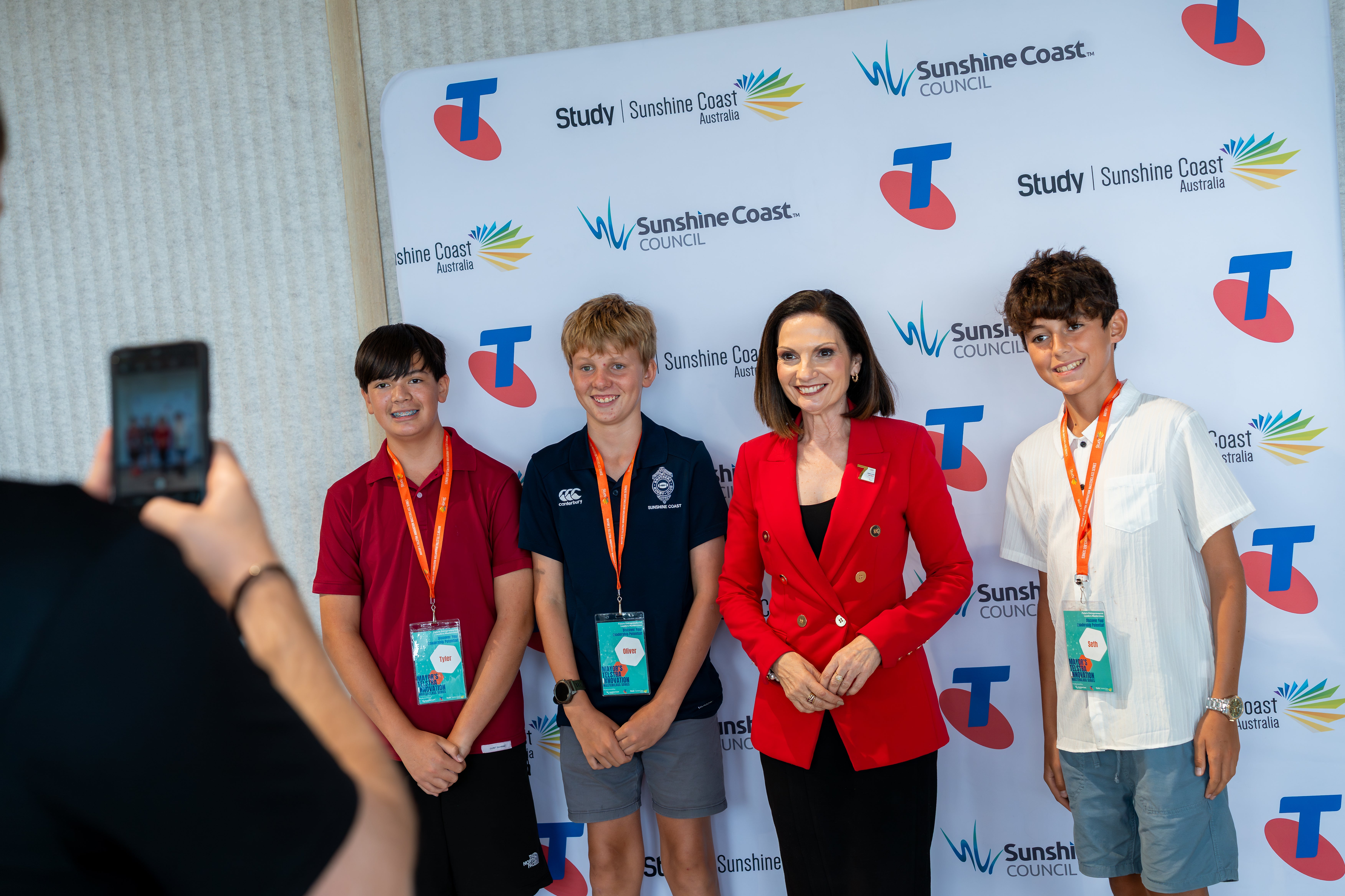 An image of someone taking a photo (in the left corner) of three students with Mayor Natoli (in a red jacket), standing in front of the sponsors board with logos for Sunshine Coast Council, Study Sunshine Coast and Telstra
