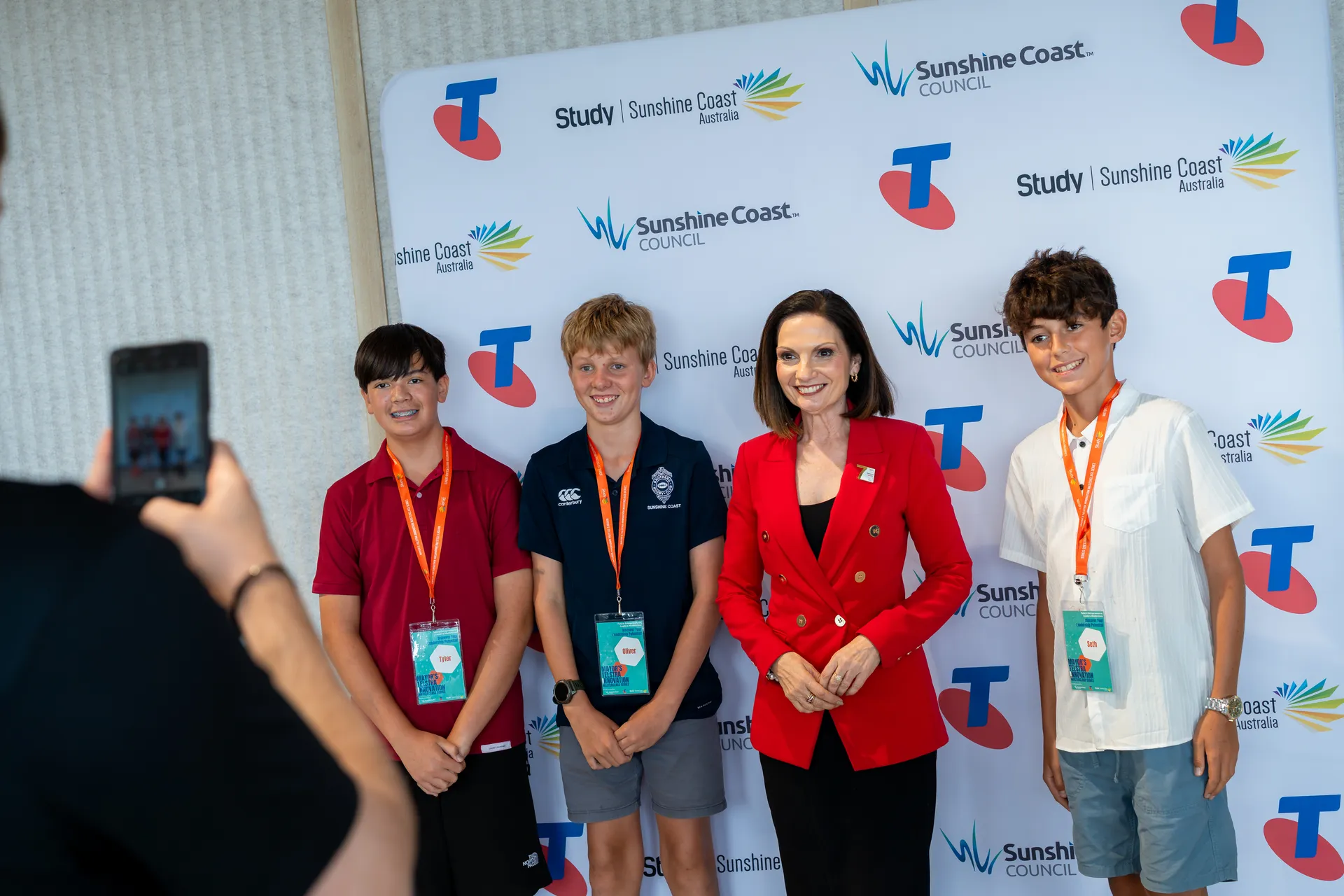 An image of someone taking a photo (in the left corner) of three students with Mayor Natoli (in a red jacket), standing in front of the sponsors board with logos for Sunshine Coast Council, Study Sunshine Coast and Telstra