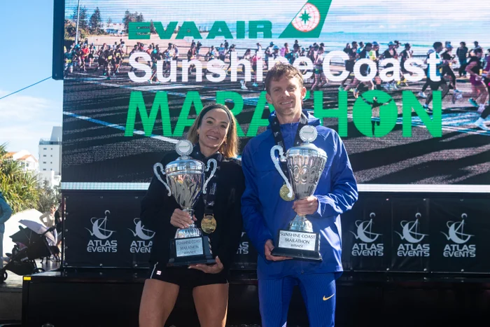 American Beth McKenzie (pictured above)and two-time Olympian Brett Robinson have claimed the marathon wins at the 2026 EVA Air Sunshine Coast Marathon Festival. Holding their trophies with the big screen behind them showing the event name EVA AIR Sunshine Coast Marathon.