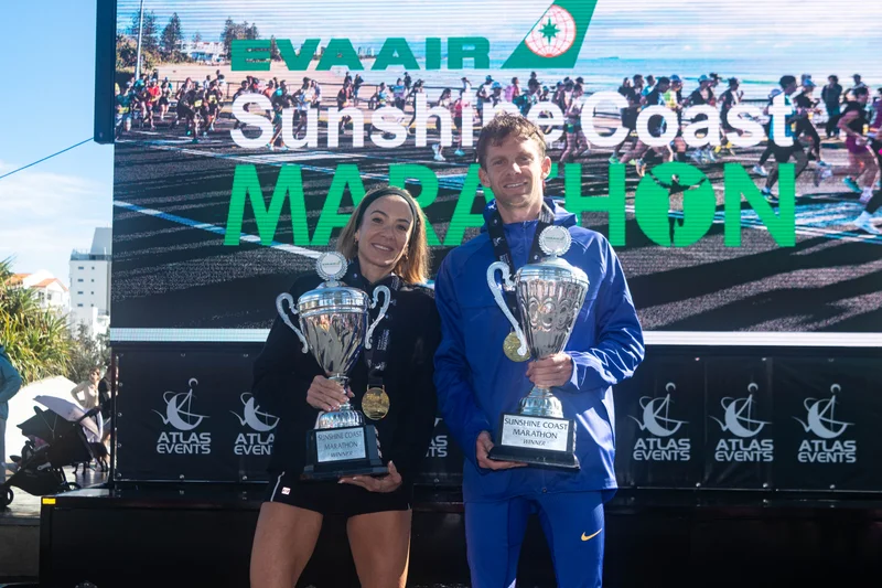American Beth McKenzie (pictured above)and two-time Olympian Brett Robinson have claimed the marathon wins at the 2026 EVA Air Sunshine Coast Marathon Festival. Holding their trophies with the big screen behind them showing the event name EVA AIR Sunshine Coast Marathon.