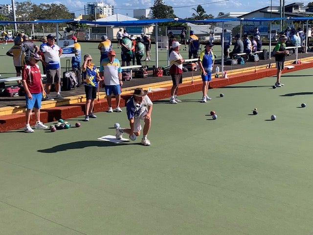 Set on a bowling green, one person almost kneeling, with other bowlers standing behind, and other bowls scattered  to the right