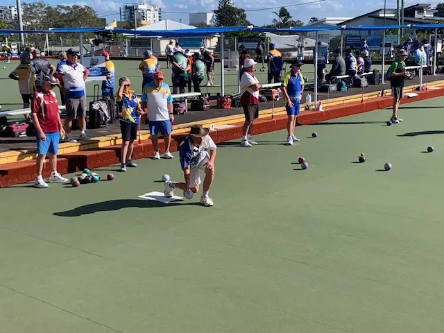 Set on a bowling green, one person almost kneeling, with other bowlers standing behind, and other bowls scattered to the right