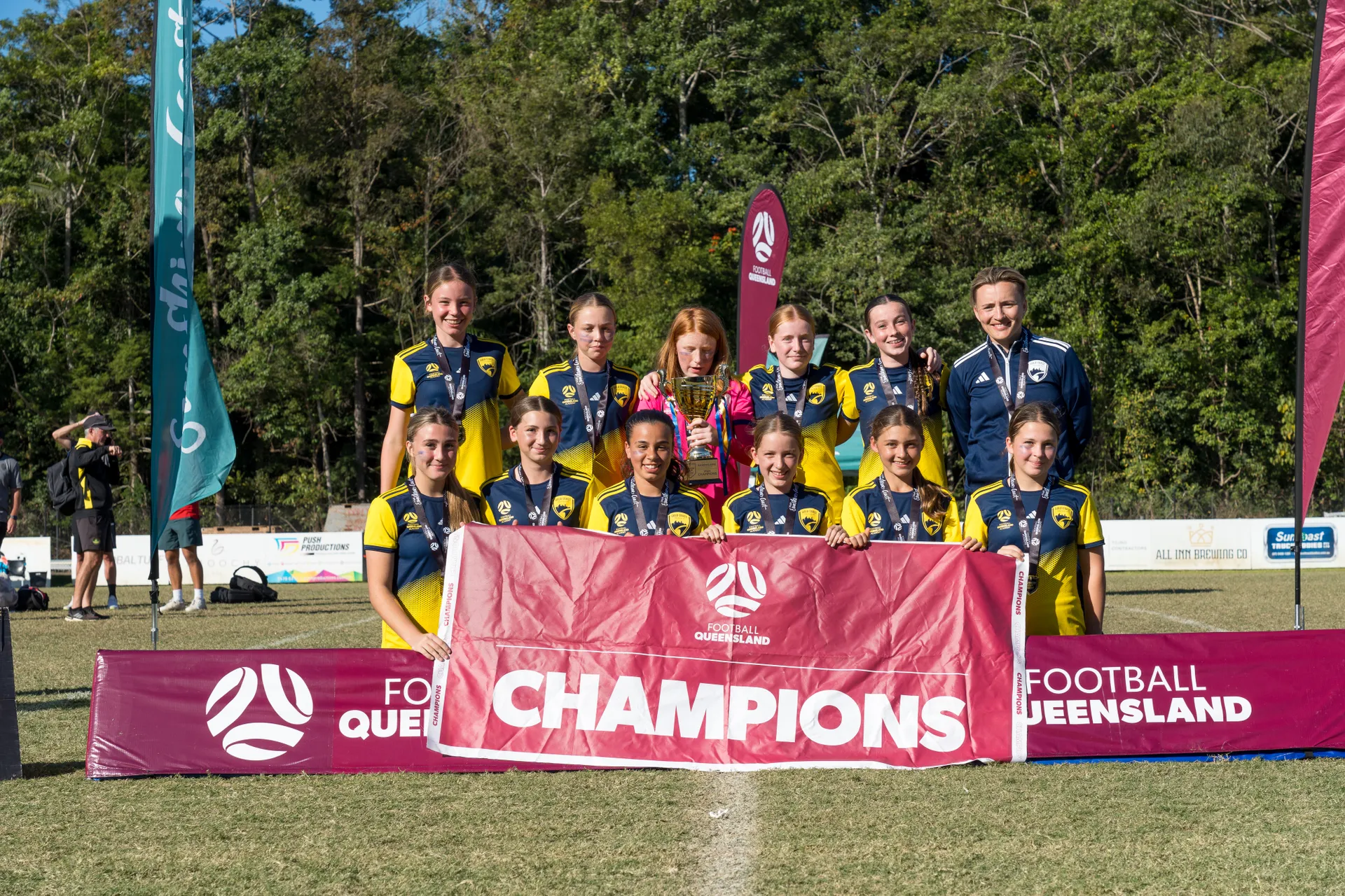 2 levels of 11 players dressed in green and yellow (gold) and coach in the green colour far right - holding up a maroon coloured Champions banner.