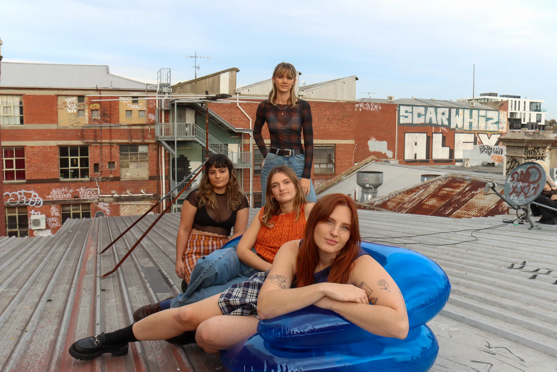 Betty Taylor Band - 4 band members - one standing, one kneeling behind a blue inflatable lounge chair with two seated on the inflatable - it appears this is set up on a rooftop with old buildings in the background with graffiti.