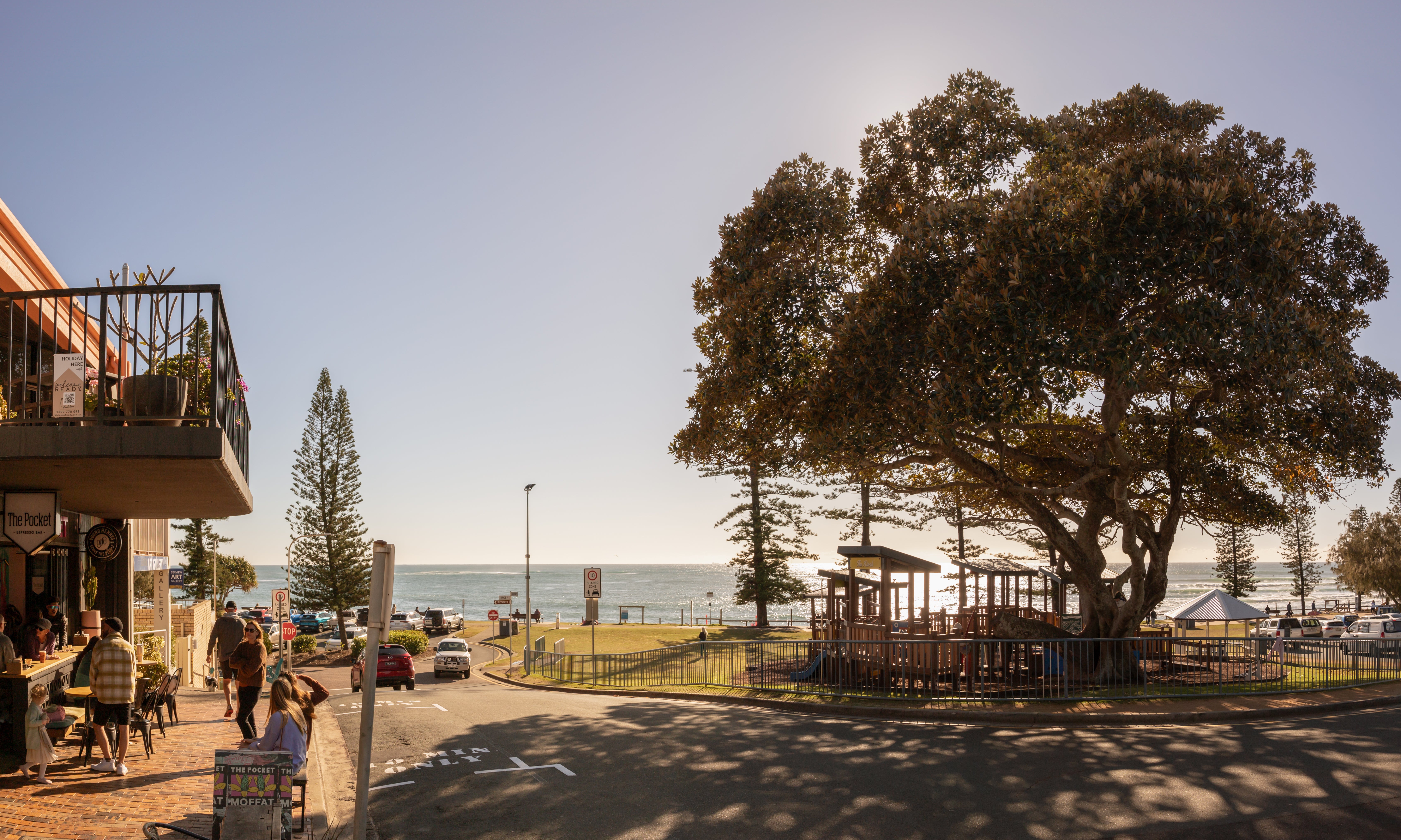 A lively playground with a large tree providing shade overhead. The ocean can be seen in the background, while shops line the left side. People are walking and enjoying life, with a spacious green area adding to the scene.