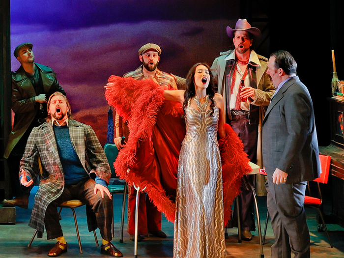 Woman in a gold dress and a red feathery coat singing opera. Surrounded by gentlemen in smart suits singing with her. 