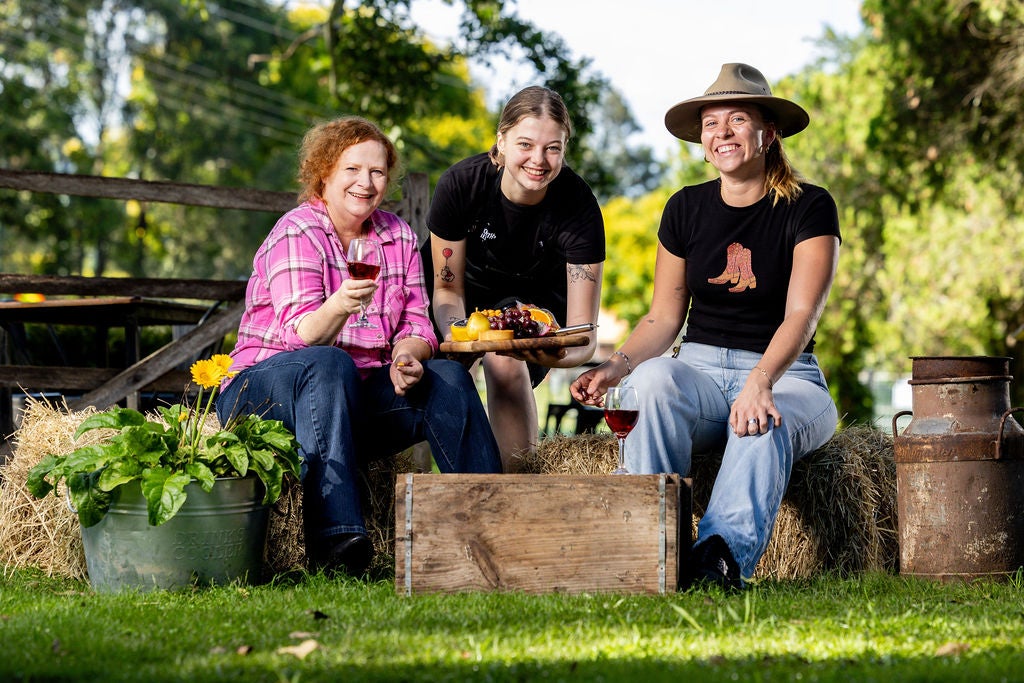 Three people sitting on hay bales - one holding a glass of wine, one presenting a charchuterie board