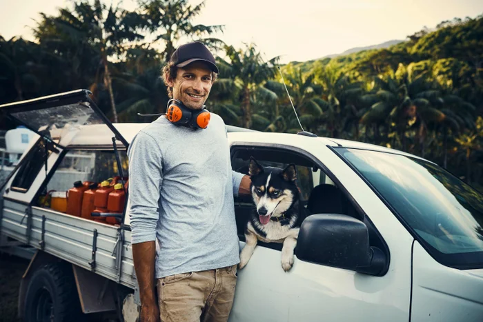 A man stands in front of a trade ute with a dog on the front seat.