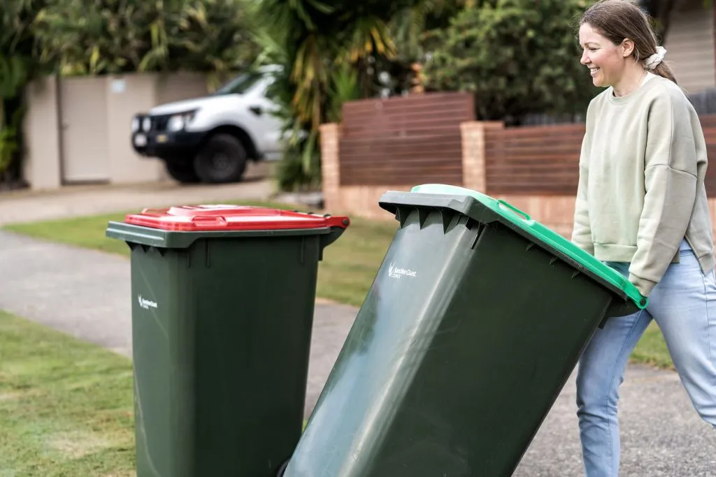 On a residential street, a lady is placing a green-lidded bin on the nature strip.