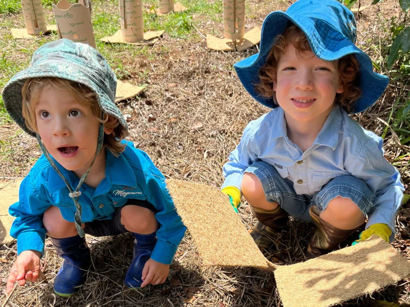 Two boys crouch, one holding plant matting material.