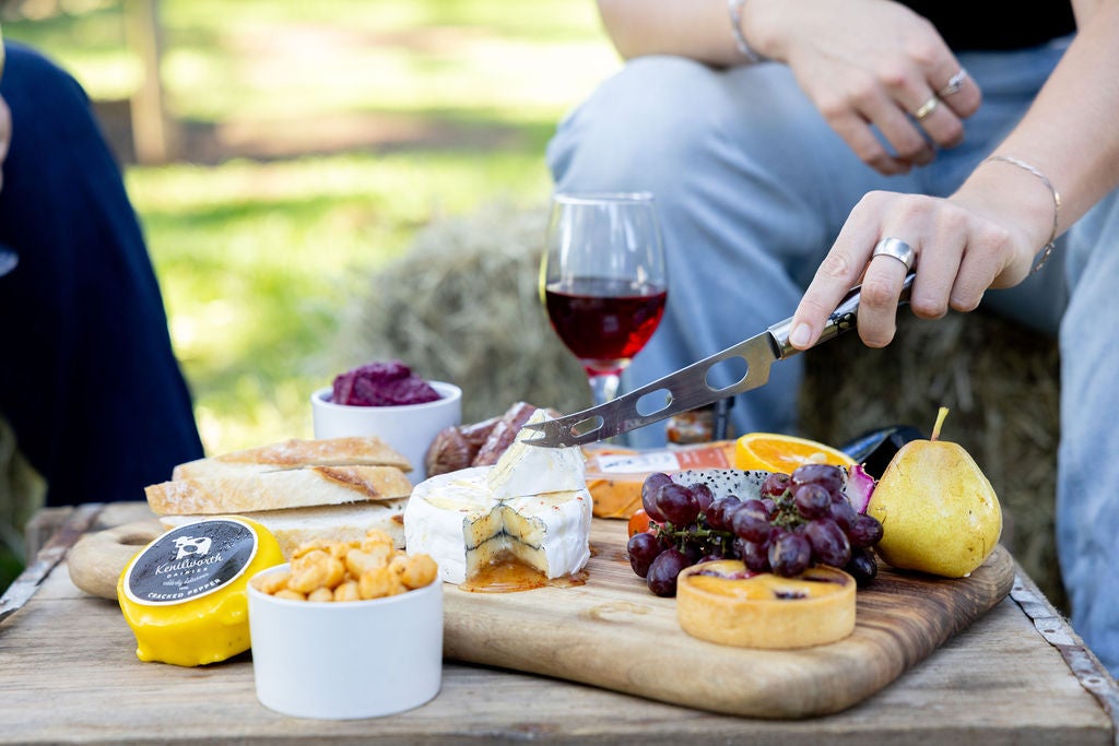 A chopping board filled with cheeses, grapes, a pear and more, a glass of wine behind, and a person's hand holding a cheese knife about to cut into a cheese.